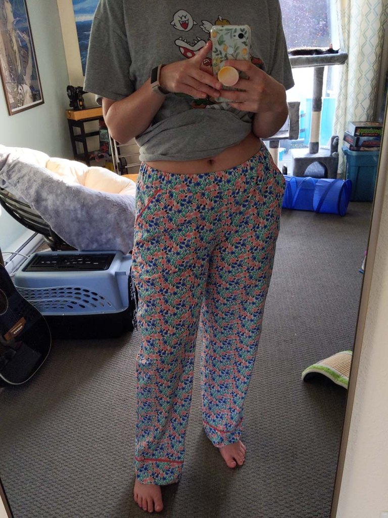 A mirror selfie of Roxanne, a woman in her early 30s. She is wearing a pair of floral Carolyn PJ pants. Her cat Owen is sunbathing in the background.