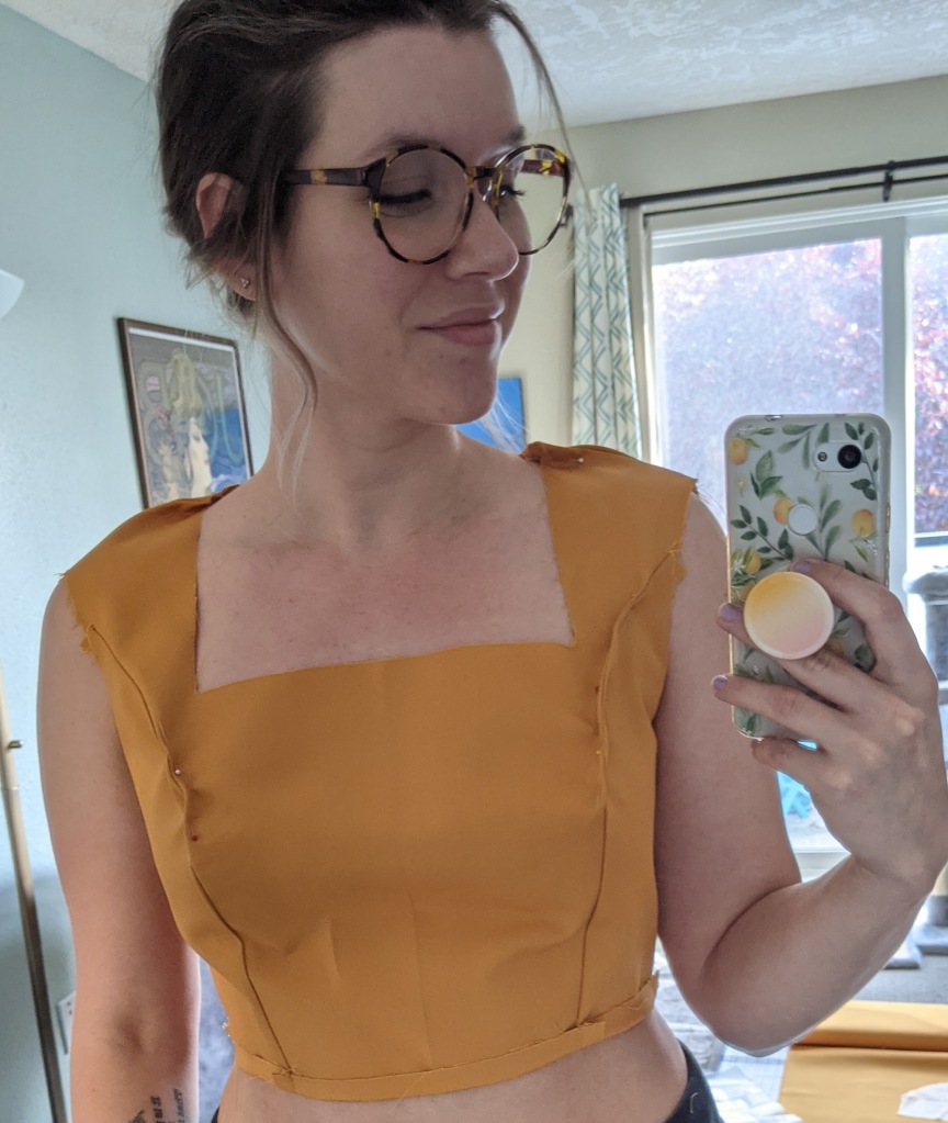 Roxanne is taking a mirror selfie in a muslin of the McCall's M6887 bodice. It is made of a yellow fabric and is pinned above the shoulders and bust.